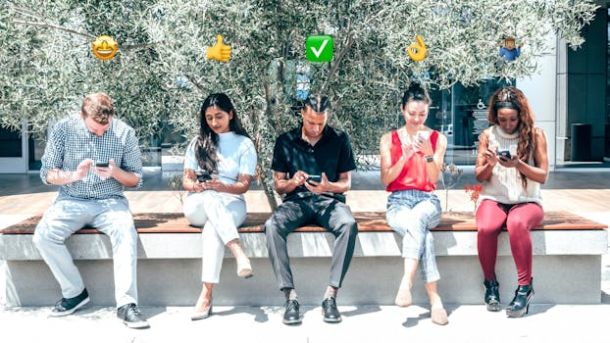Diverse group of people sitting outdoors using smartphones under emojis.